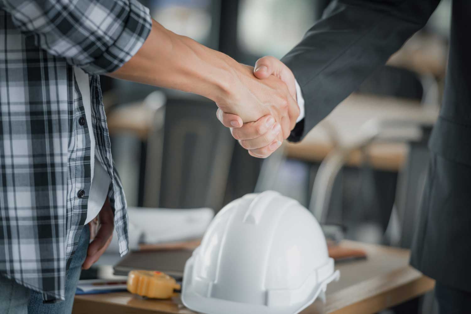 Handshake between two individuals in a business setting, with a construction helmet and tools on the table, symbolizing collaboration and partnership in technology and IT services.